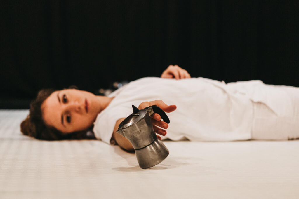 Una donna si lascia andare sul pavimento, in preda alla depressione. In una mano stringe una piccola caffettiera, che osserva con sguardo vuoto.