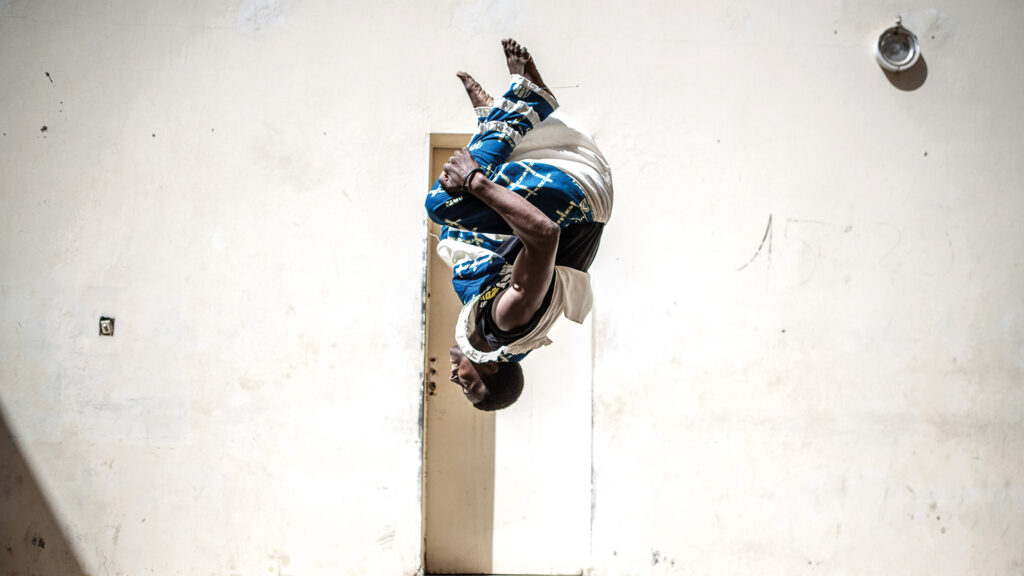 Un uomo senegalese in costume tradizionale esegue un salto mortale all'indietro. Come Together 2025
