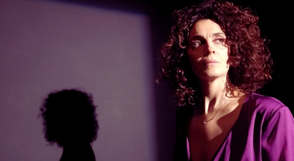 una donna riccia guarda davanti a sé, sul muro la sua ombra